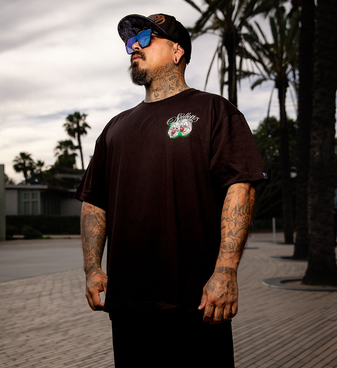 Man wearing a black t-shirt with a logo, sunglasses, and cap outdoors.