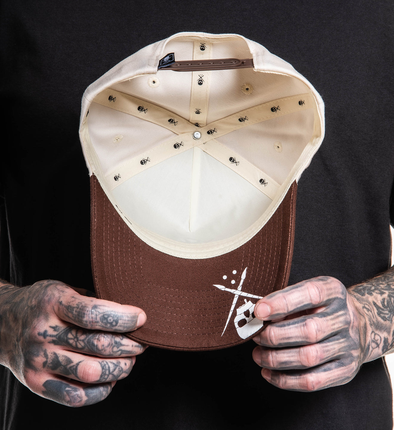 Brown and beige cap held by a person with tattoos against a black background