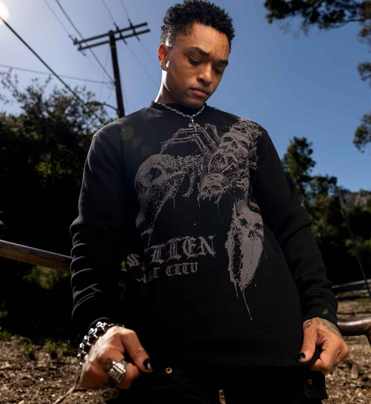 A man wearing a black long-sleeve graphic sweatshirt with a large gray skull and crossbones design and the text 'SAXION THE CROW' on the front, standing outdoors against a blue sky and trees.