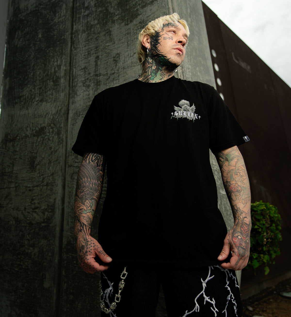 Person wearing a black t-shirt with a logo, standing against a textured wall.