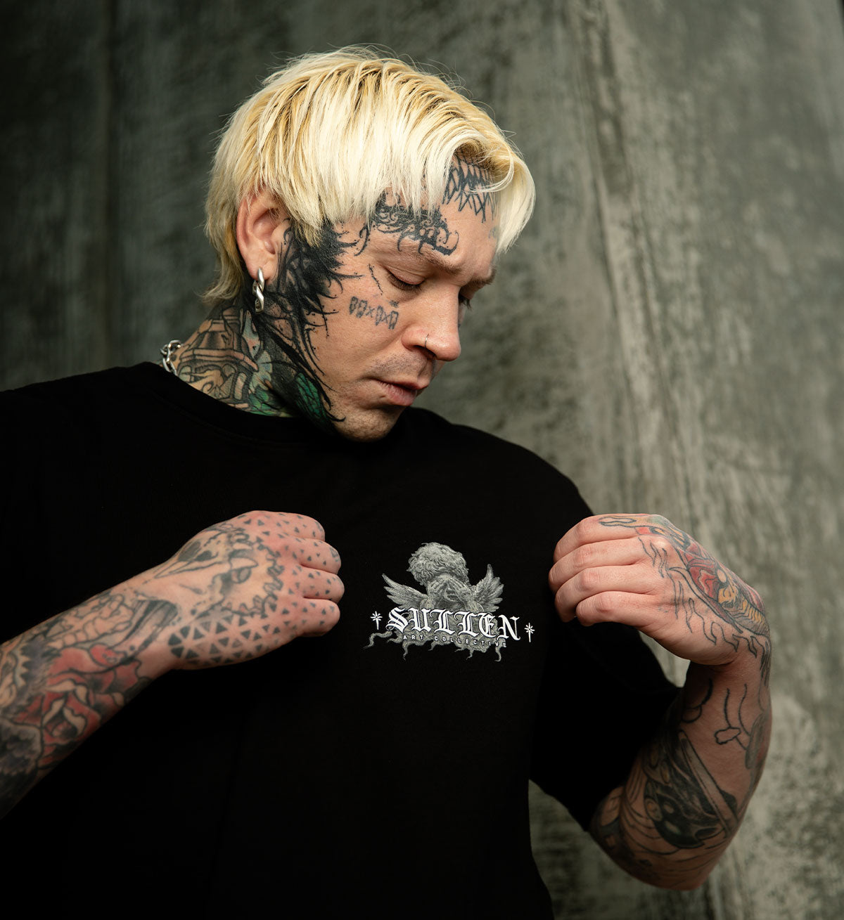 Person wearing a black t-shirt with a logo, standing against a textured gray background