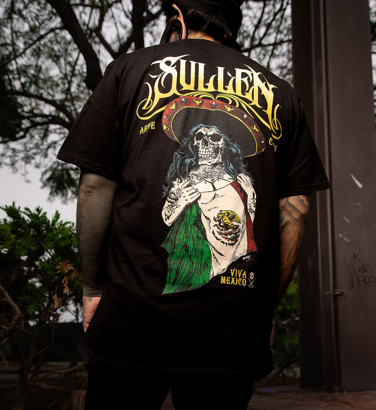 Person wearing a black t-shirt with a graphic design on the back, featuring a skeleton in a sombrero and green dress.