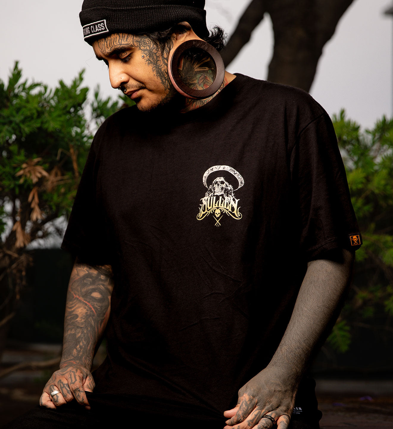 Man wearing a black t-shirt with a logo, sitting outdoors with greenery in the background