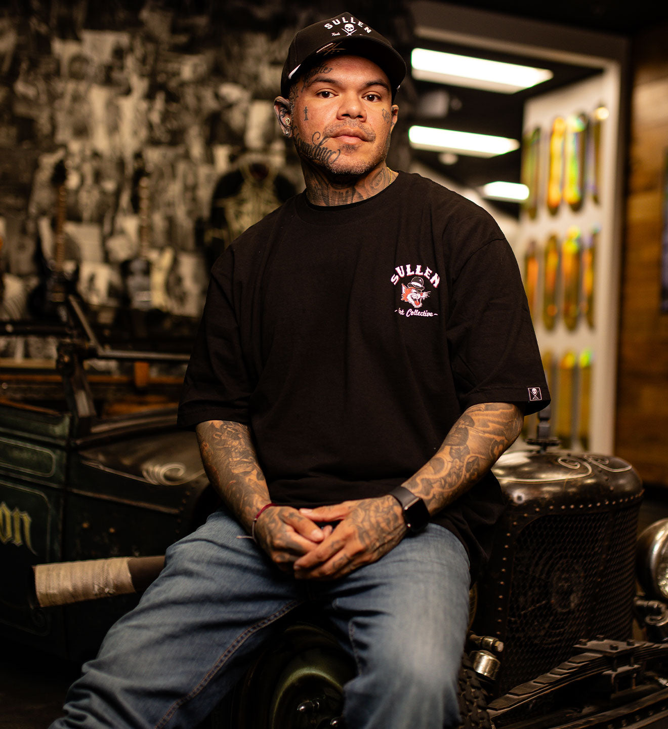 Man wearing a black t-shirt with a logo, sitting in a workshop.