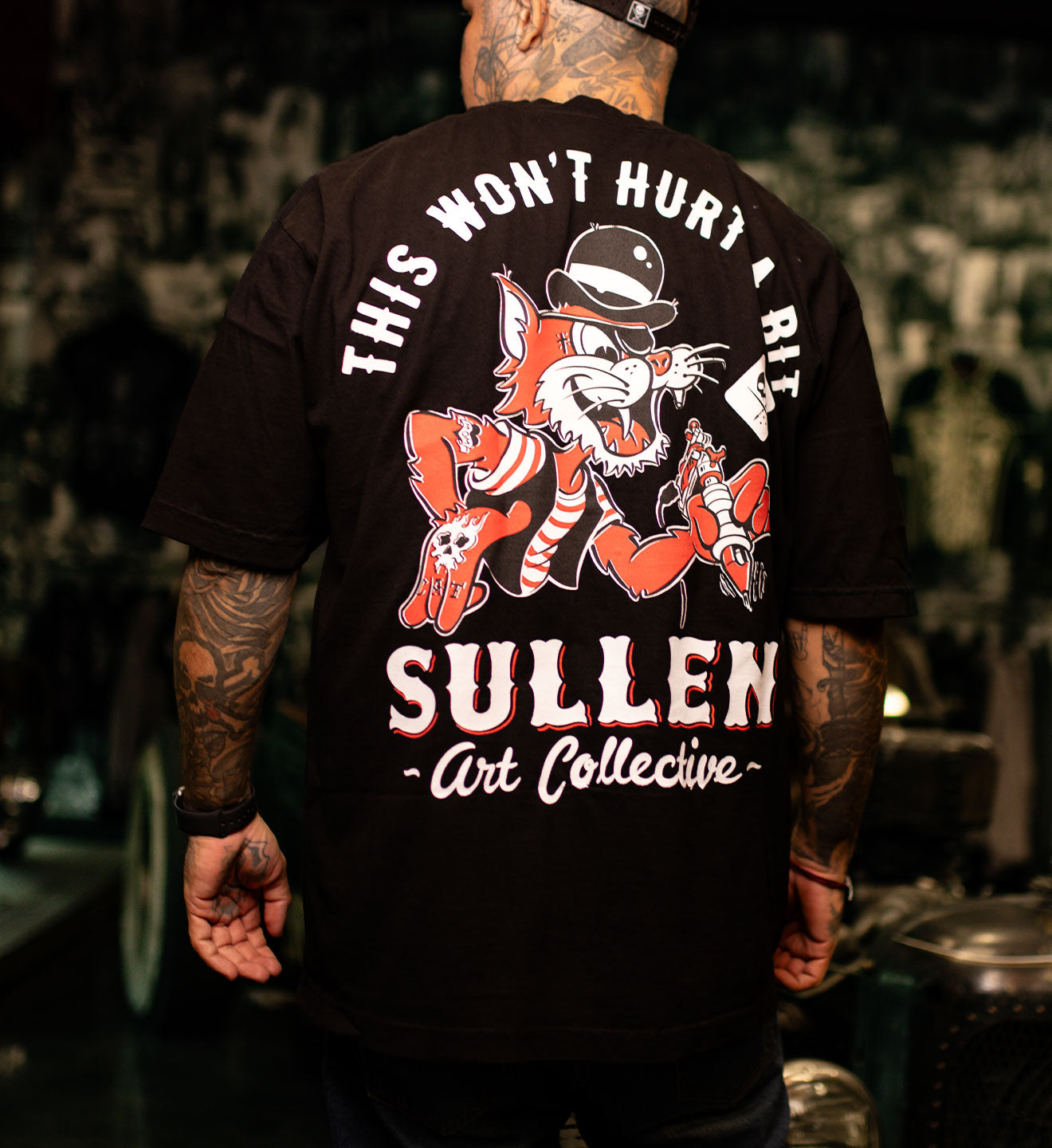 Person wearing a black t-shirt with a graphic design and text on the back, set against a blurred background.