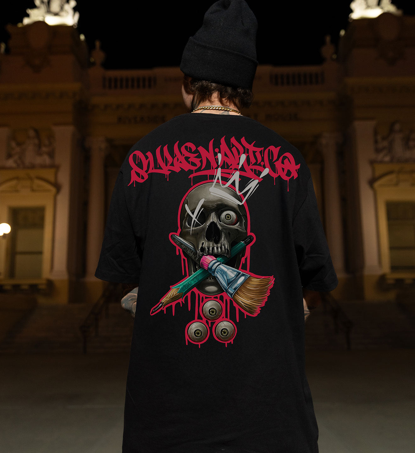 Person wearing a black t-shirt with a graphic design and text, standing in front of a building.