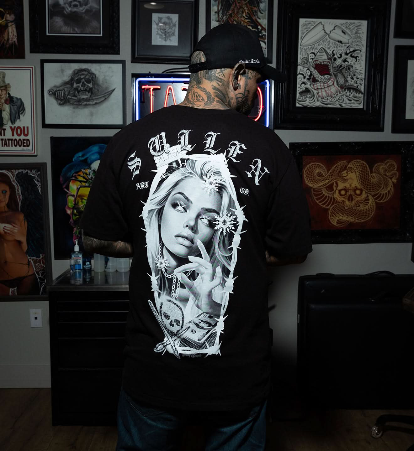 Person wearing a black t-shirt with a graphic design of a face in a coffin, standing in a tattoo parlor.