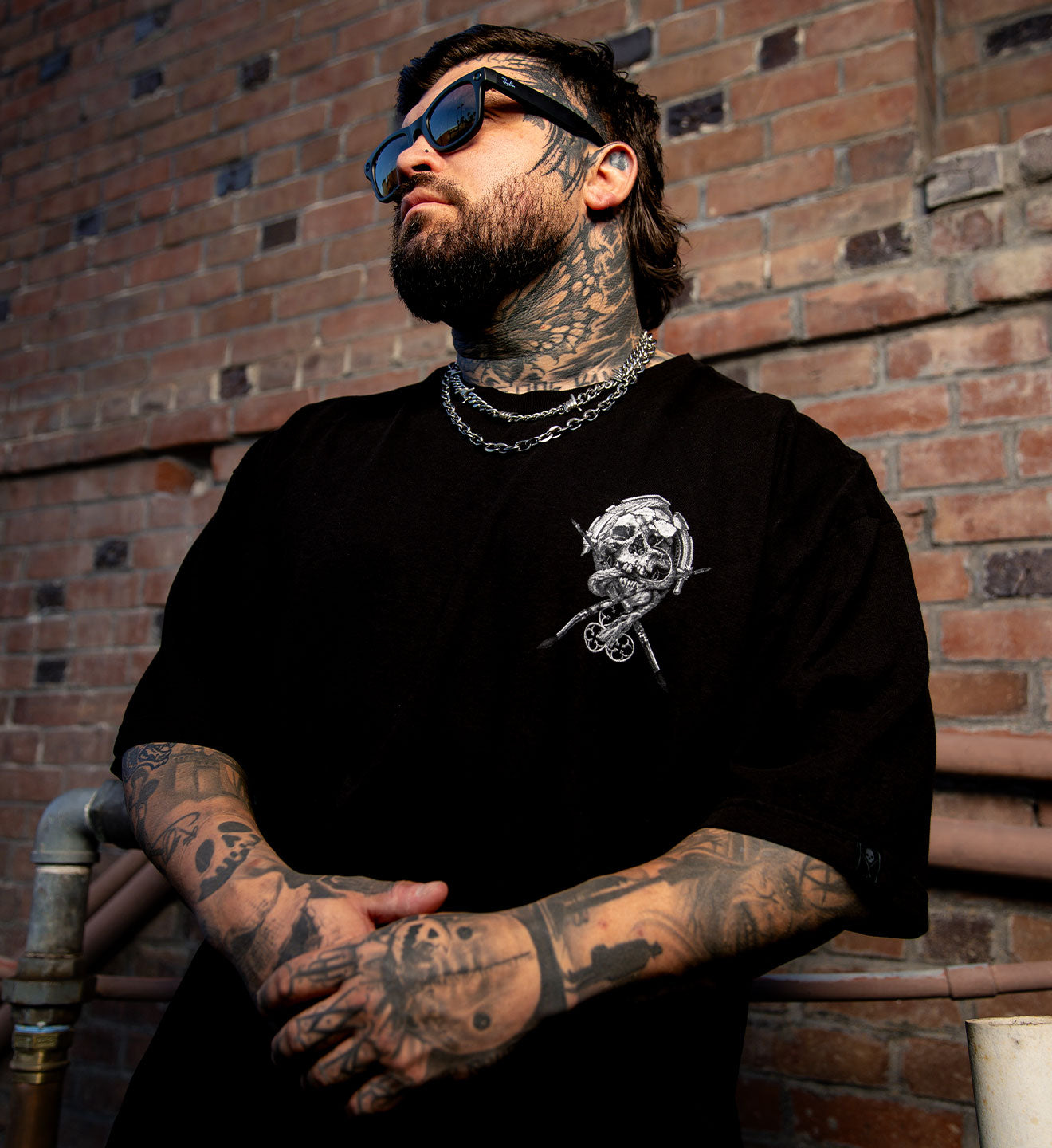 Man with tattoos wearing a black t-shirt with a skull design against a brick wall.