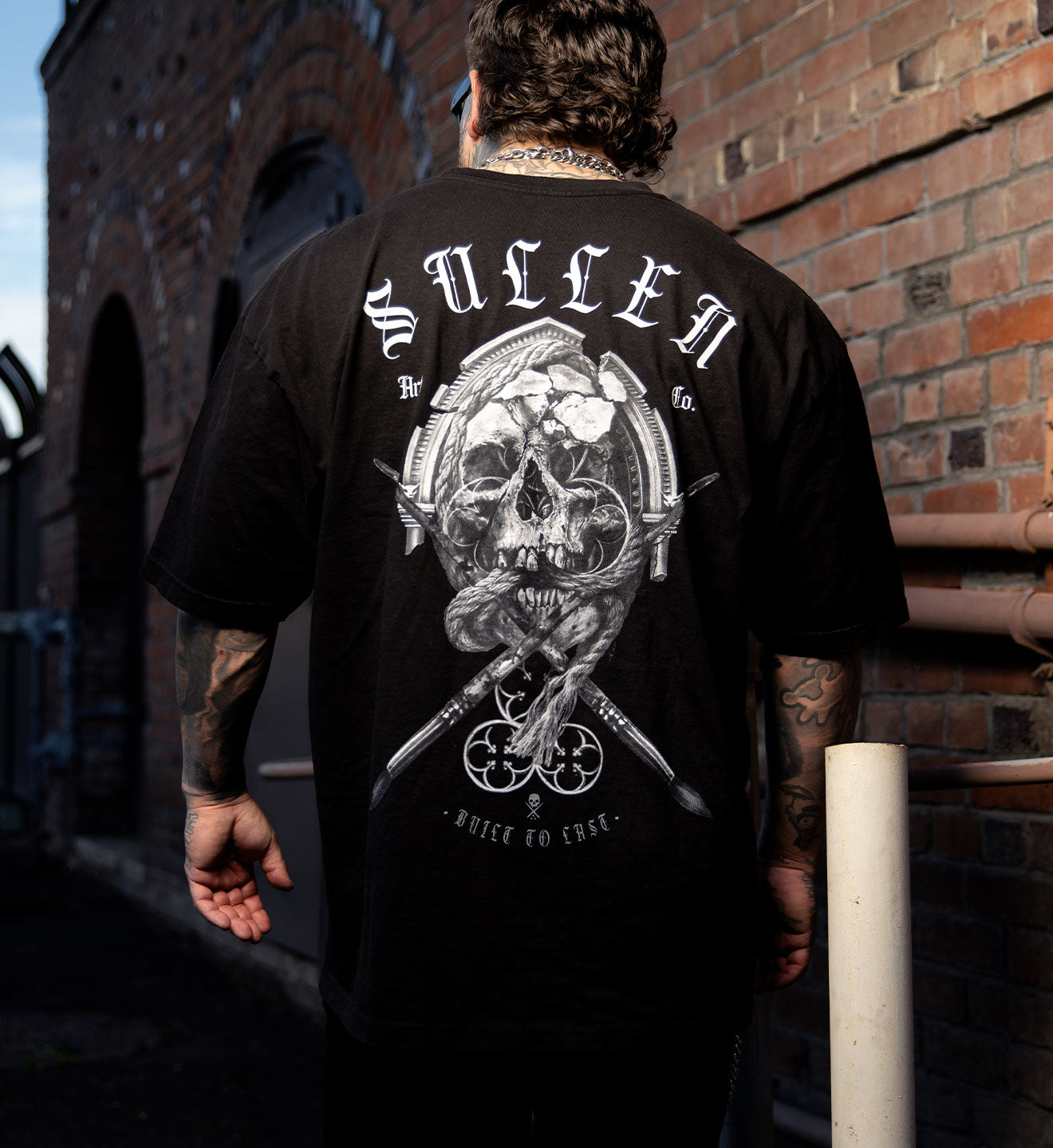 Person wearing a black t-shirt with a skull design and text, standing against a brick wall.