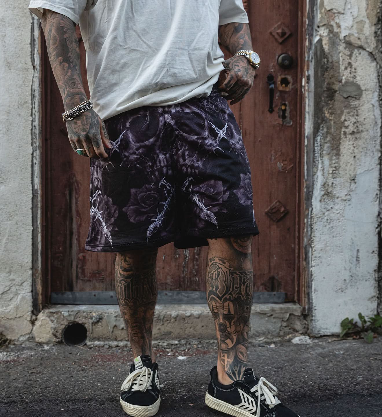 Person wearing a white t-shirt, purple patterned shorts, and black sneakers with tattoos on arms and legs, standing in front of a rustic door.
