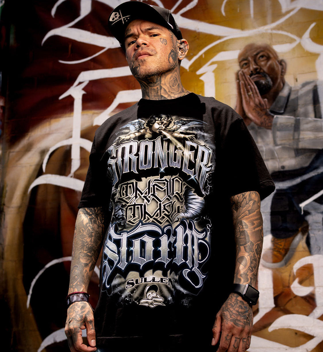 Man wearing a graphic t-shirt with text and tattoos, standing in front of a mural.
