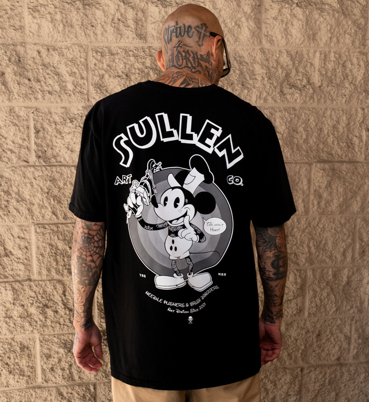 Person wearing a black t-shirt with a graphic design and 'Sullen' branding against a beige brick wall.