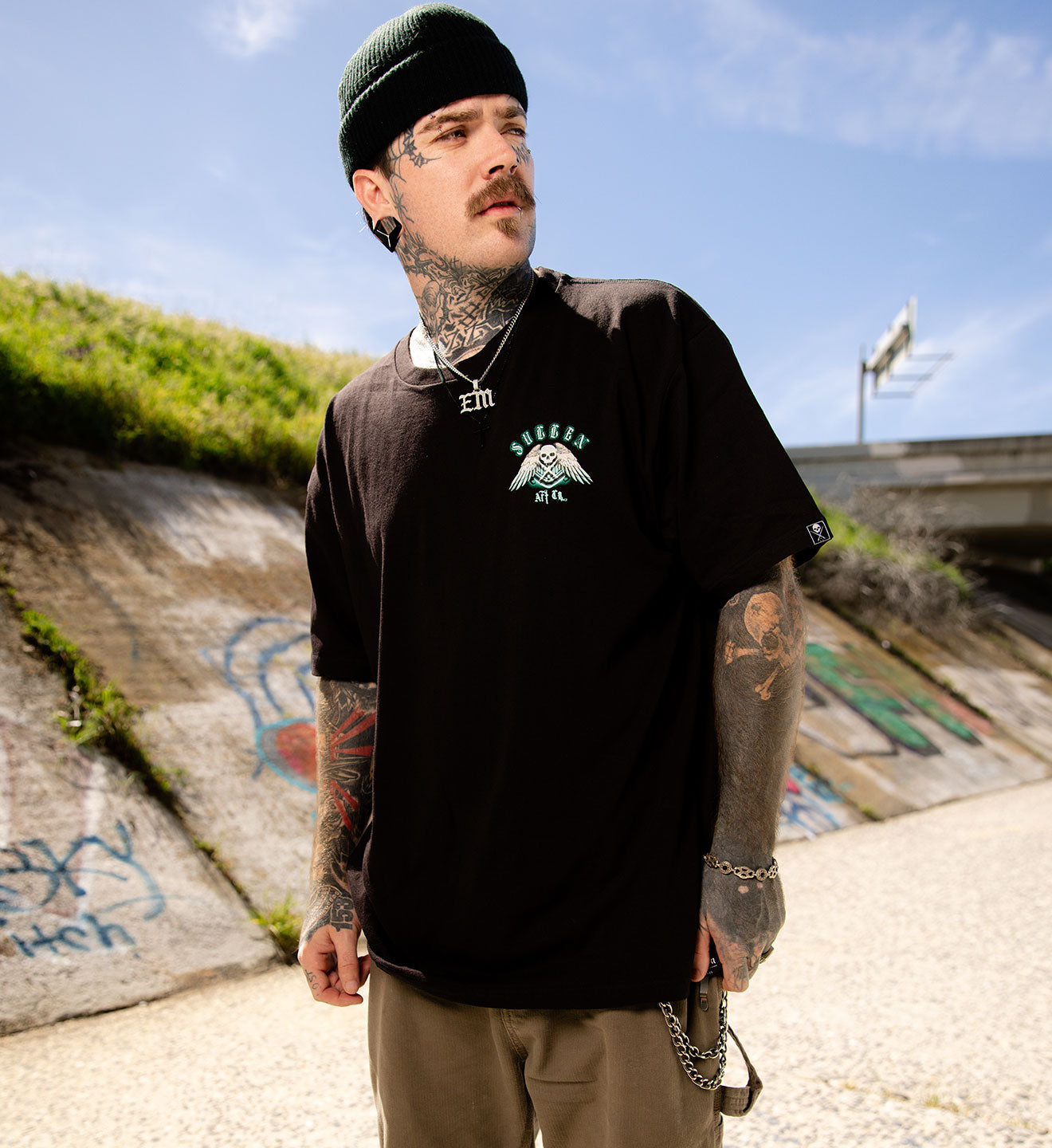 Man with tattoos wearing a black t-shirt with a logo, standing in an urban setting with graffiti.