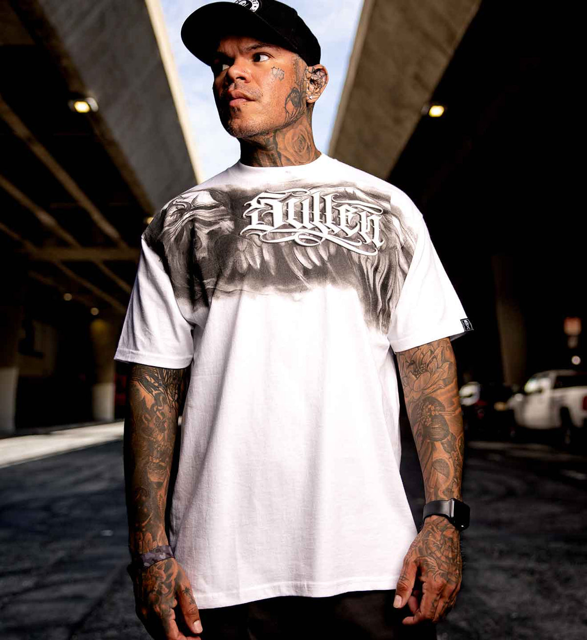 A tattooed man wearing a white graphic t-shirt with a large gray eagle design and the word 'Sullen' in gothic lettering. Resurrection collection tee from Tom Berg