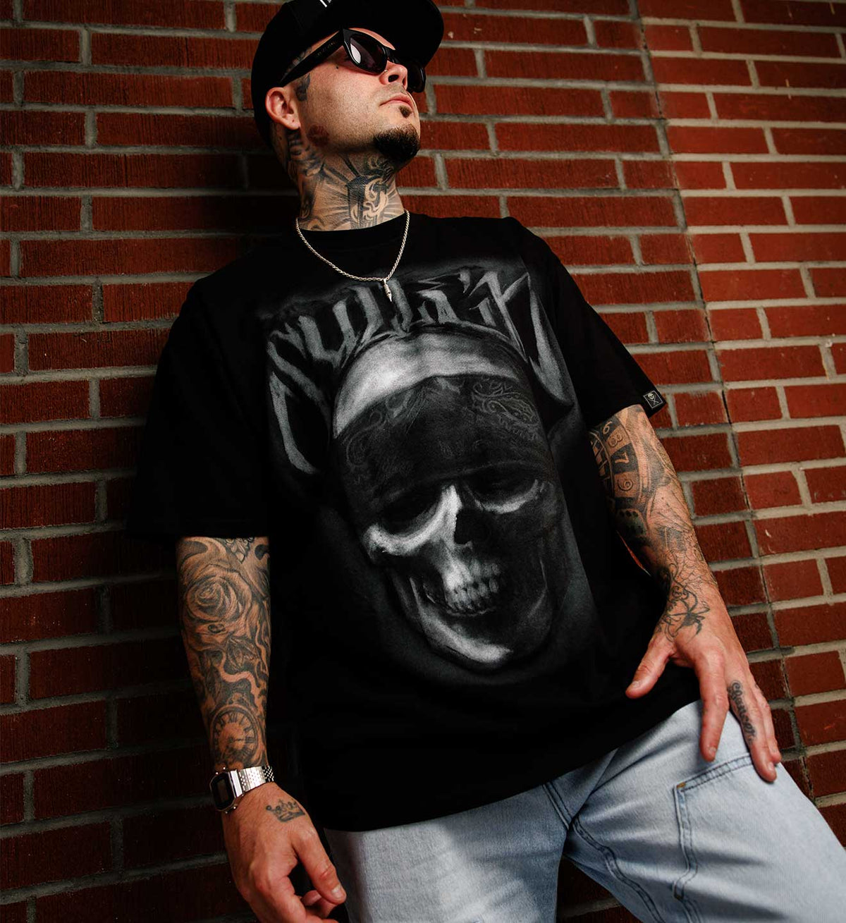 A tattooed man wearing a black band t-shirt with a large grayscale skull graphic and the word 'NUDE' in stylized lettering, paired with light blue jeans, leaning against a red brick wall.