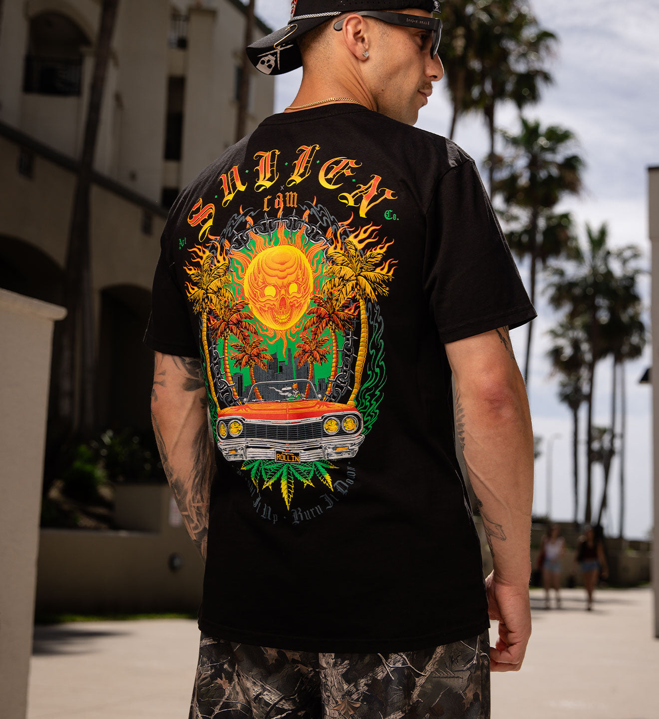 Man wearing a black t-shirt with colorful graphics and text, standing outdoors with palm trees in the background.