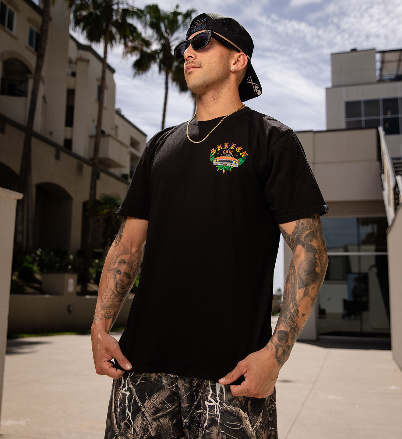 Man wearing a black t-shirt with a logo and camouflage pants standing outdoors.