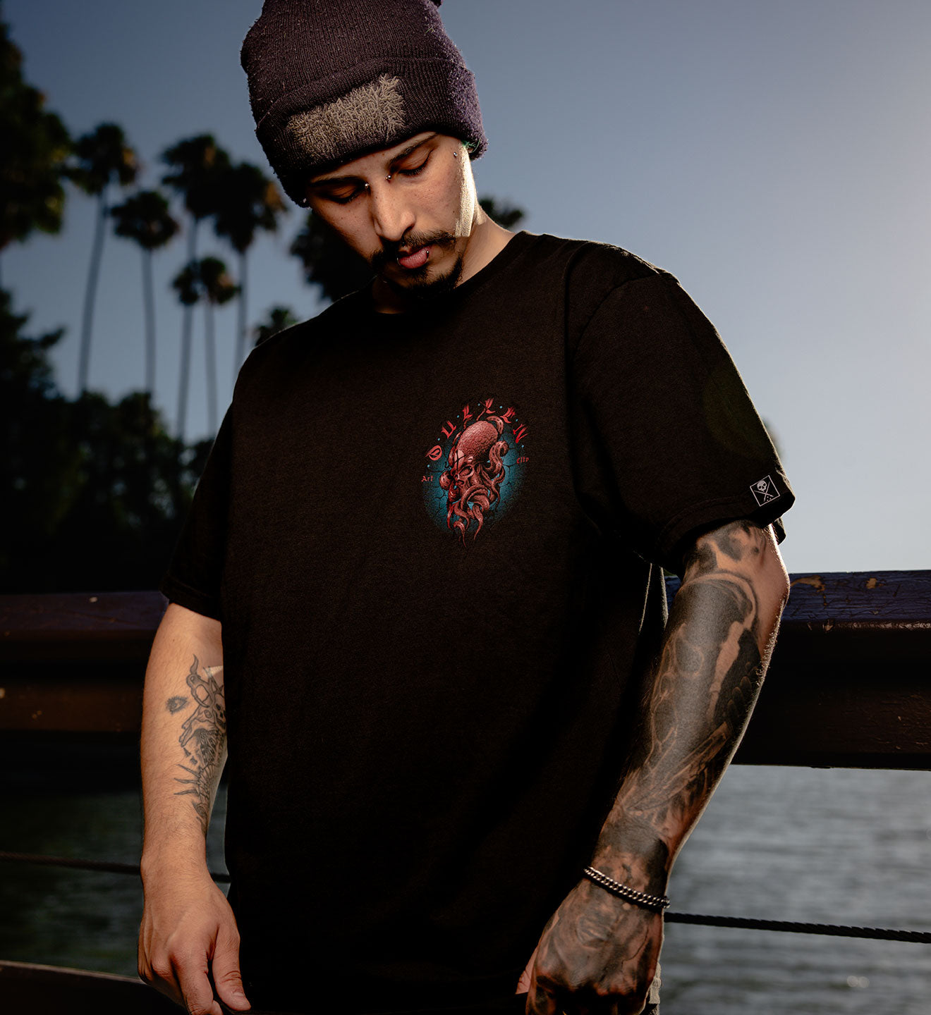 Man wearing a black t-shirt with a colorful graphic design, standing outdoors with palm trees in the background.