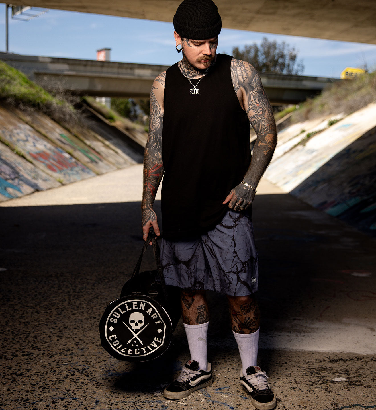 Man with tattoos holding a bag with 'Sullen Art Collective' logo in an urban setting.