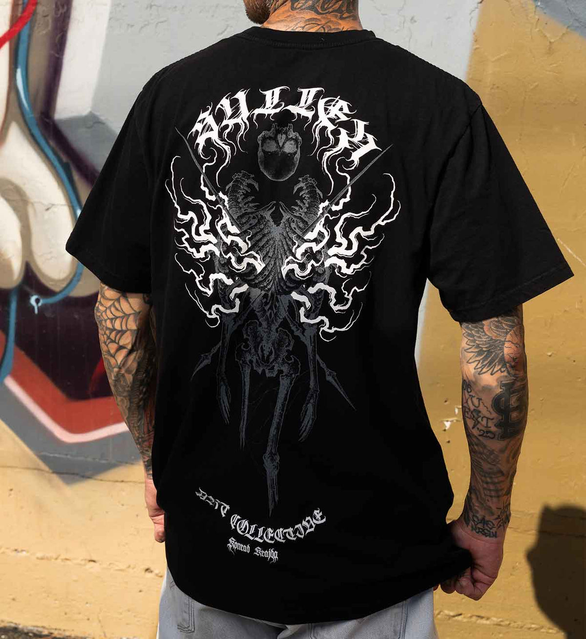 A man wearing a black t-shirt with a large white and gray graphic print on the back, standing against a graffiti-covered wall.