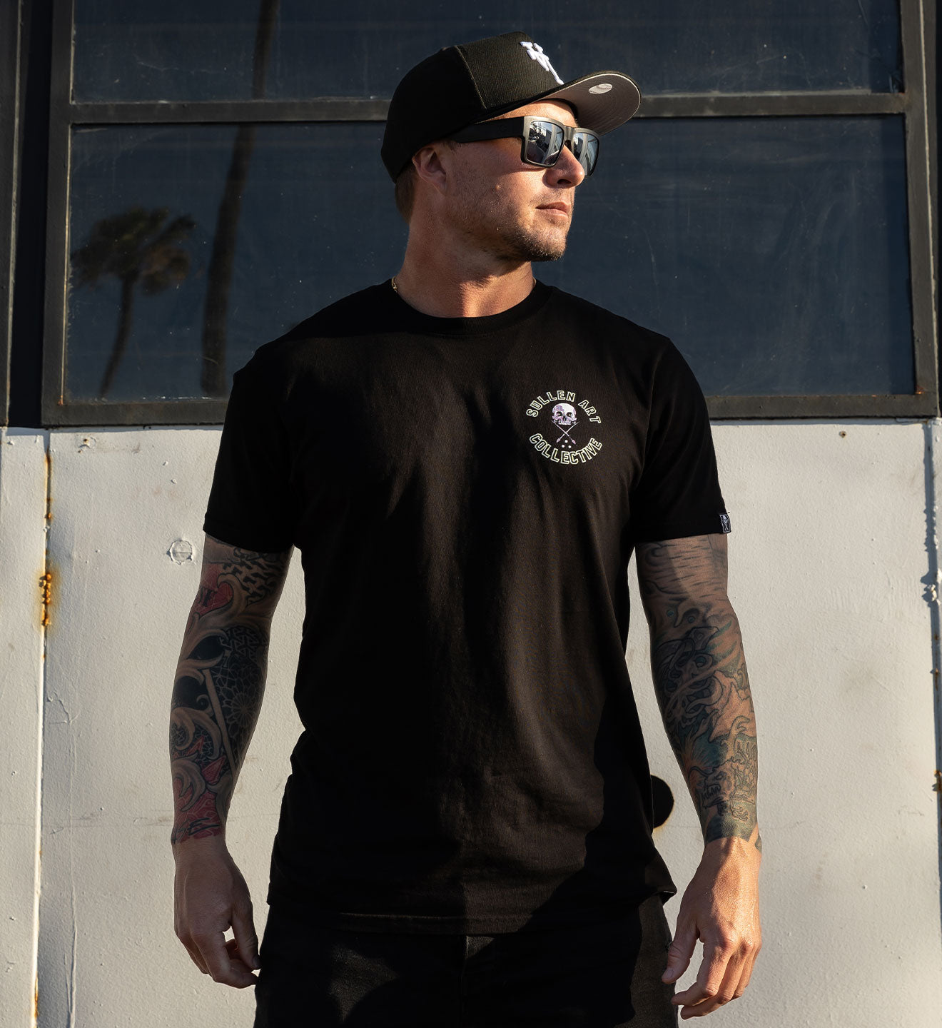 Man wearing a black t-shirt with a logo and text, standing against a wall.