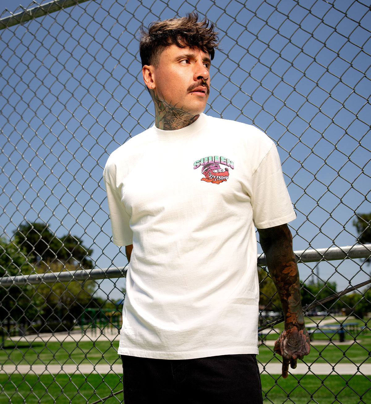 Man wearing a white t-shirt with a logo in front of a chain-link fence.