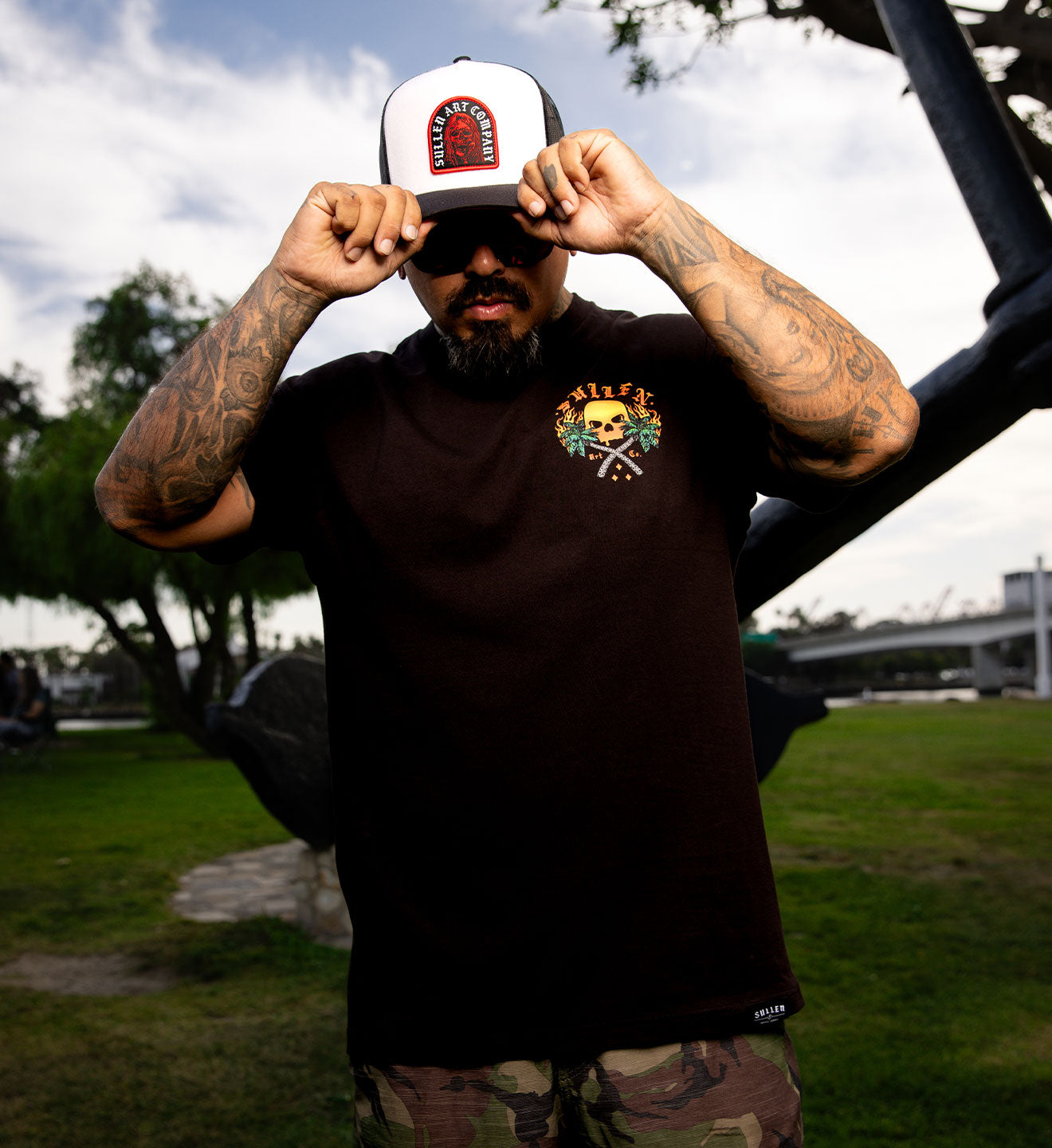 Man wearing a black t-shirt with a logo and a cap, standing outdoors.