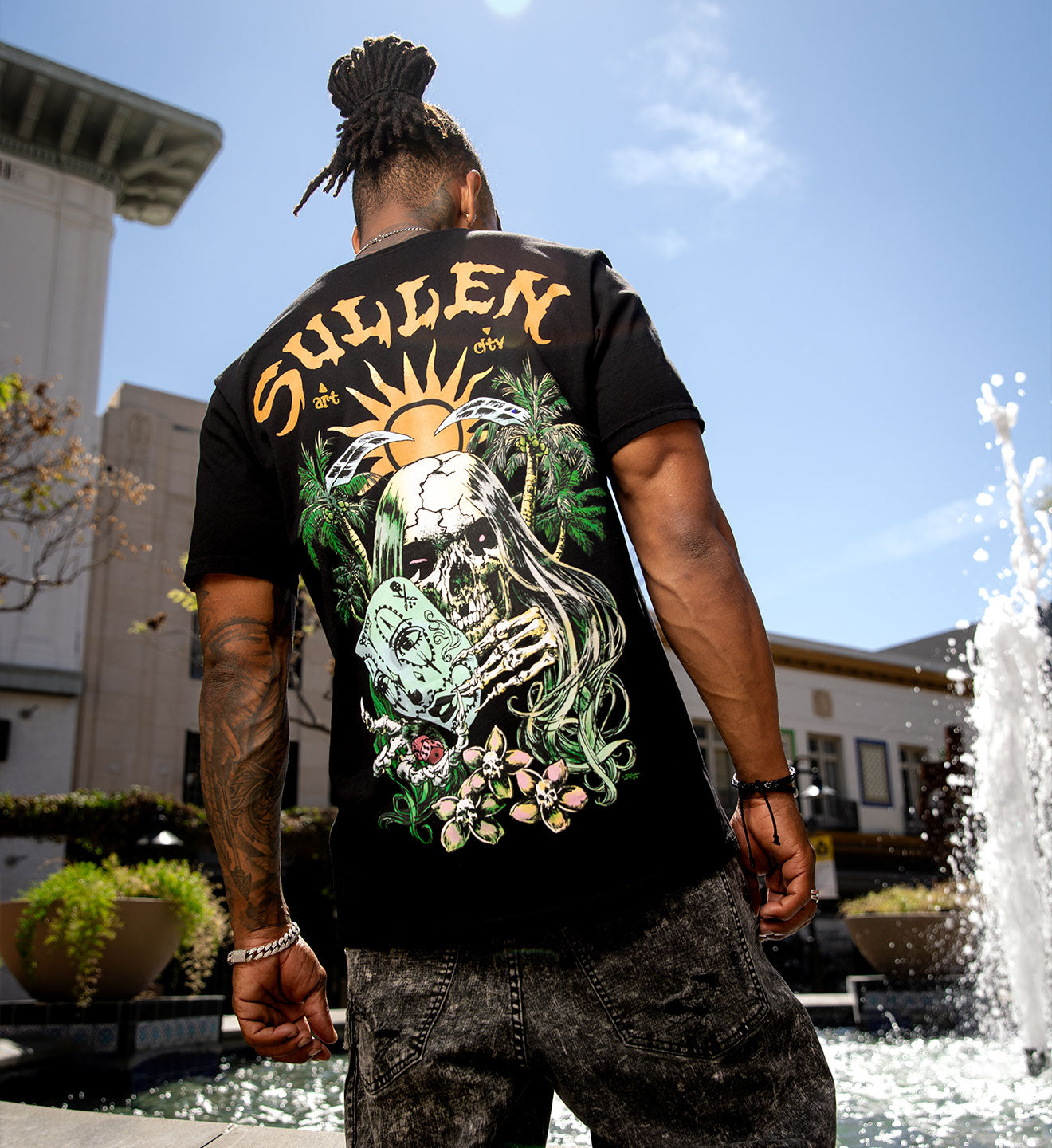 Person wearing a black t-shirt with a colorful design and 'Sullen' branding, standing outdoors near a fountain.