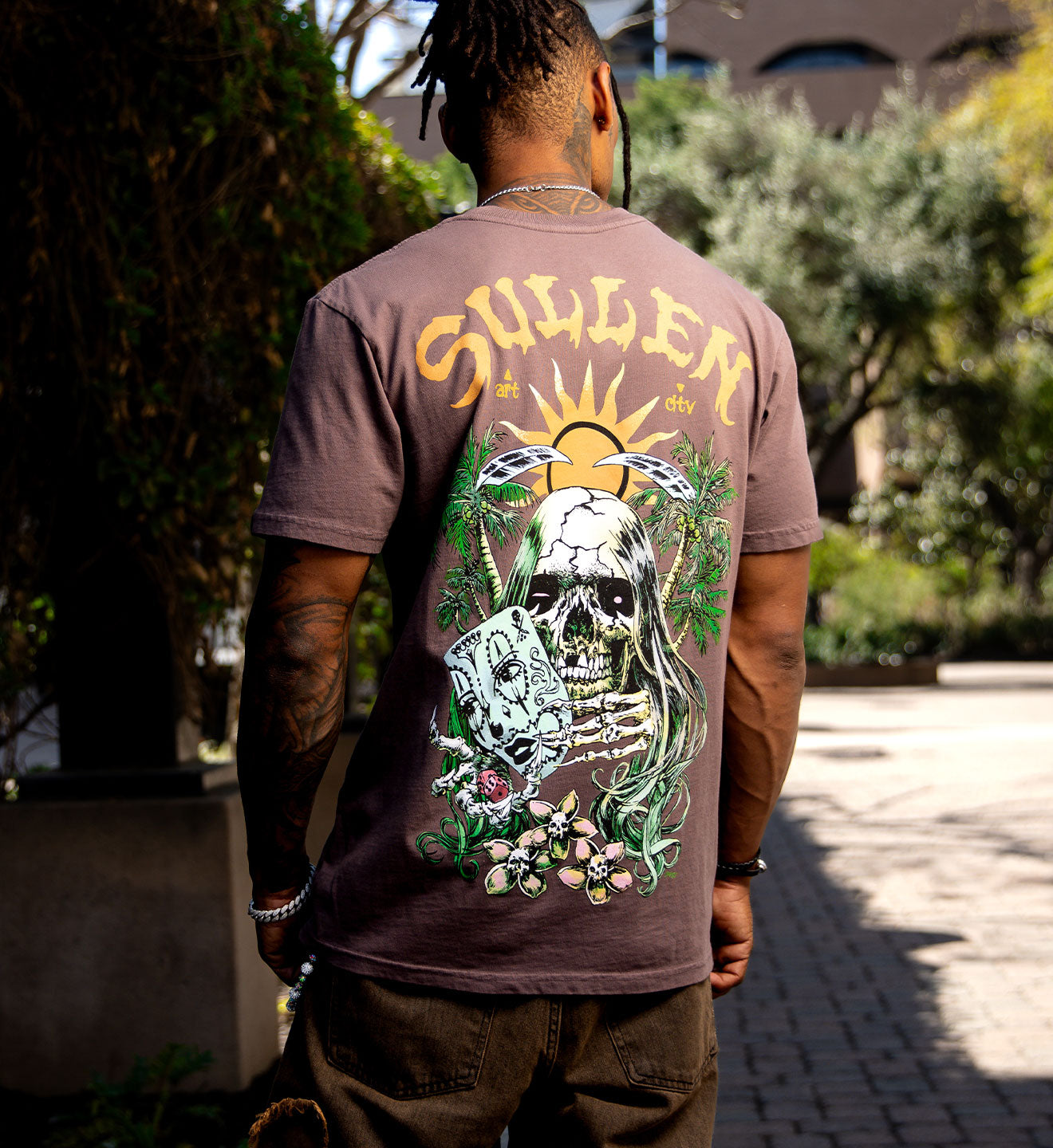 Person wearing a brown t-shirt with a graphic design on the back, standing outdoors.