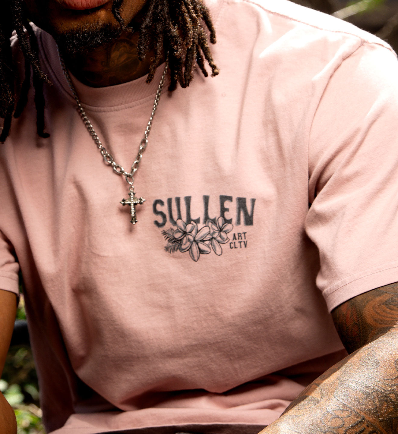 Person wearing a pink t-shirt with 'Sullen' logo and text, outdoors.