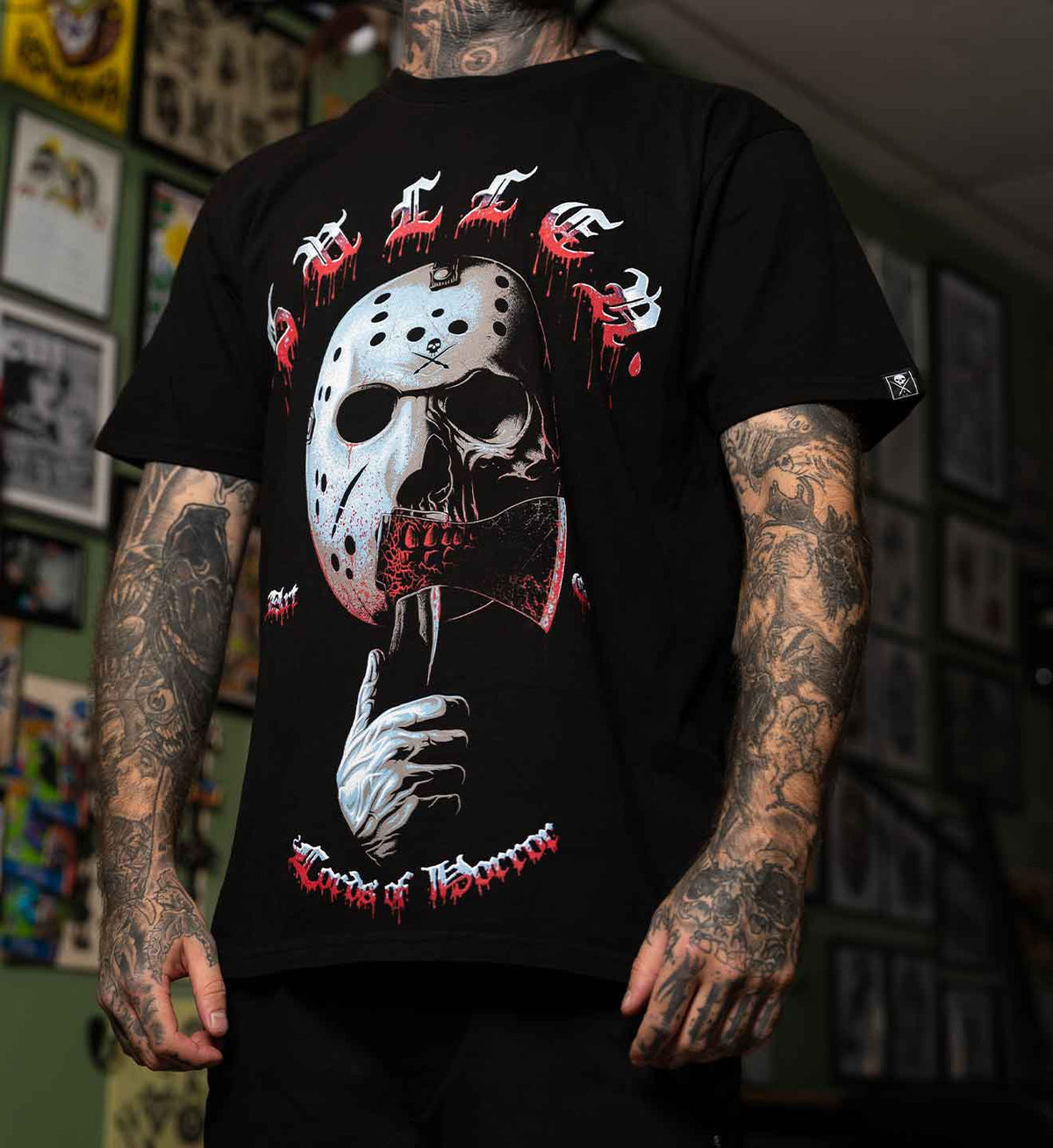 A tattooed man wearing a black graphic t-shirt featuring a horror-themed design of a hockey mask, a hand holding a bloody knife, and red dripping text that reads 'Ghosts of Halloween'.