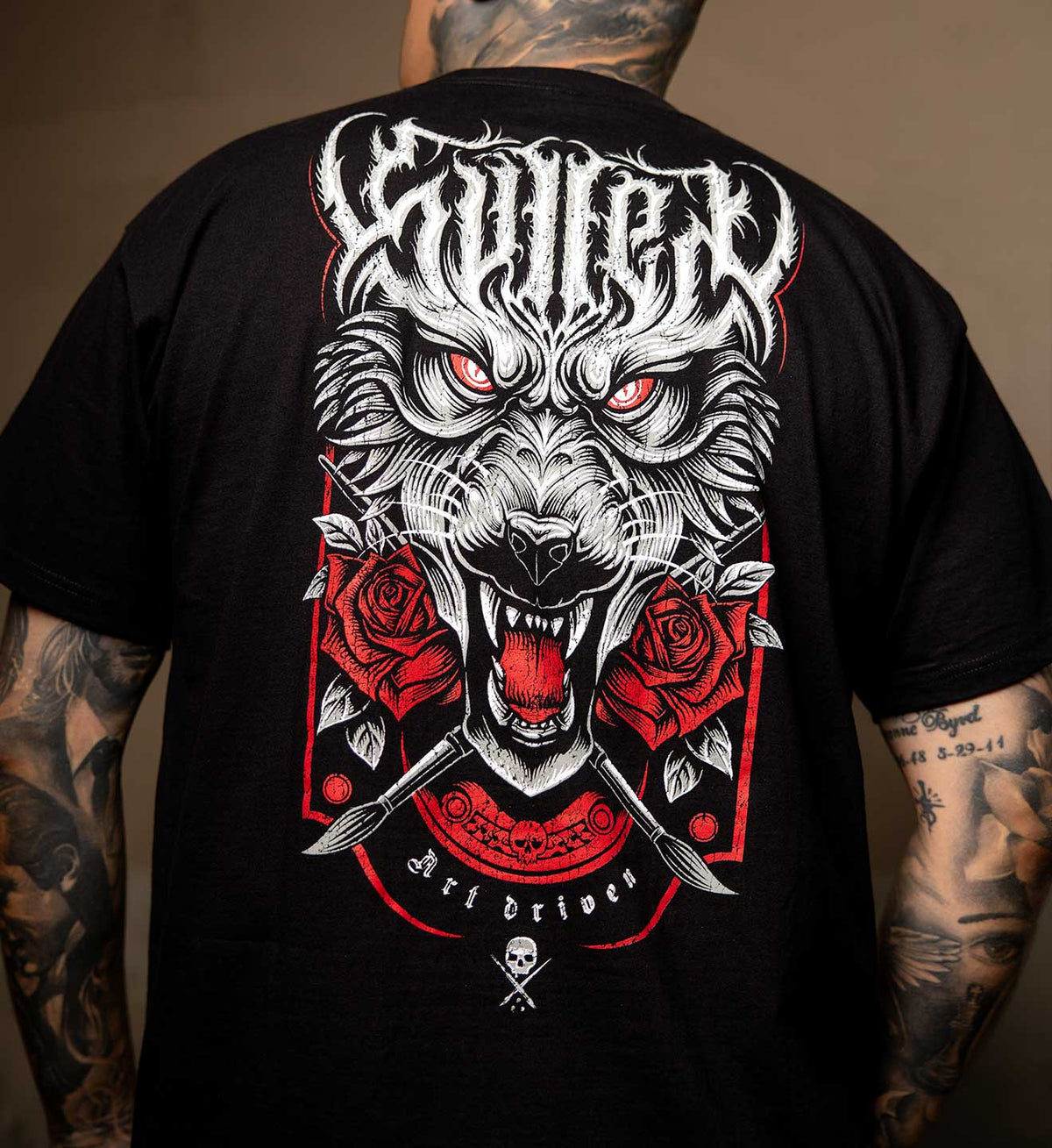 A person viewed from behind wearing a black graphic T-shirt. The shirt features a large white and red illustrated design with a snarling wolf face, red roses, crossed knives, and stylized text reading 'Sullen' and 'Art driven'.