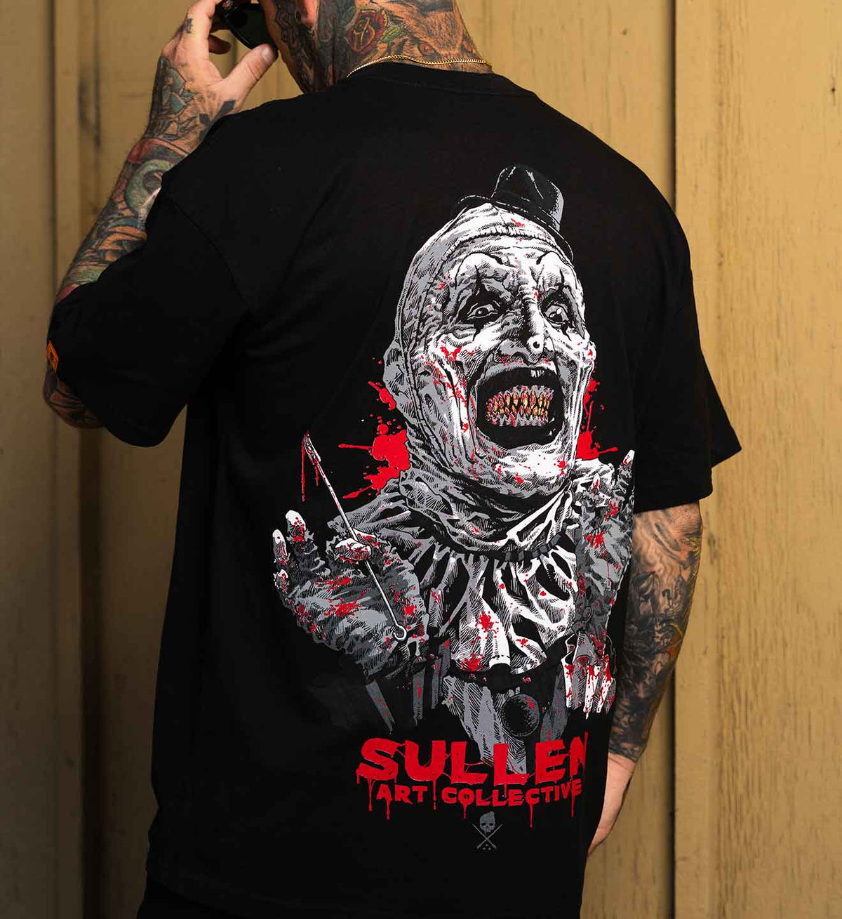 A man with tattooed arms is shown from the back wearing a black t-shirt with a large graphic print of a bloody clown and the text 'SULLEN ART COLLECTIVE' in red, set against a wooden background.
