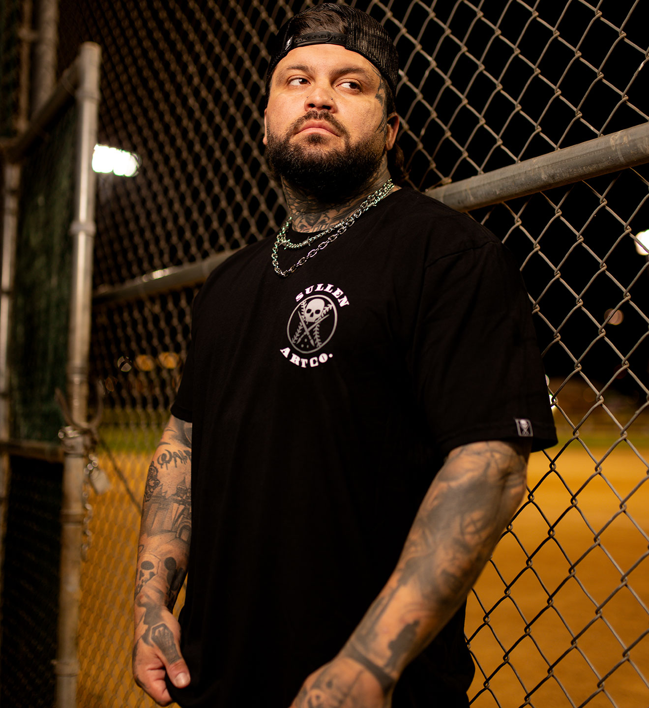 Man wearing a black t-shirt with a logo, standing in front of a chain-link fence.