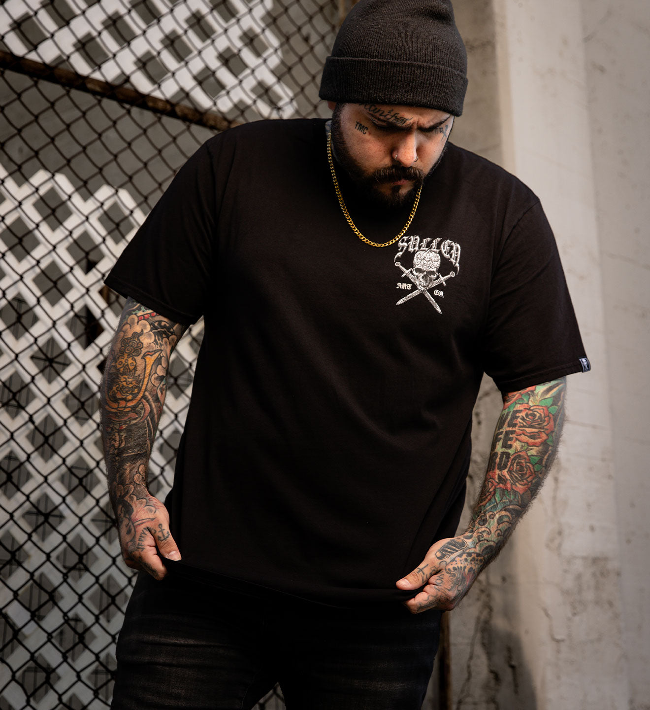 Man wearing a black t-shirt with a logo and text, standing against a chain-link fence and concrete wall.
