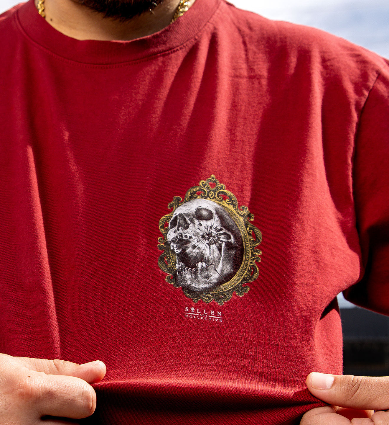 Red t-shirt with a graphic design of a skull and ornate frame on a blurred outdoor background