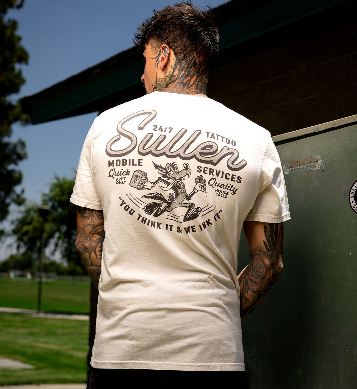 Person wearing a white t-shirt with 'Sullen' tattoo services design, standing outdoors.