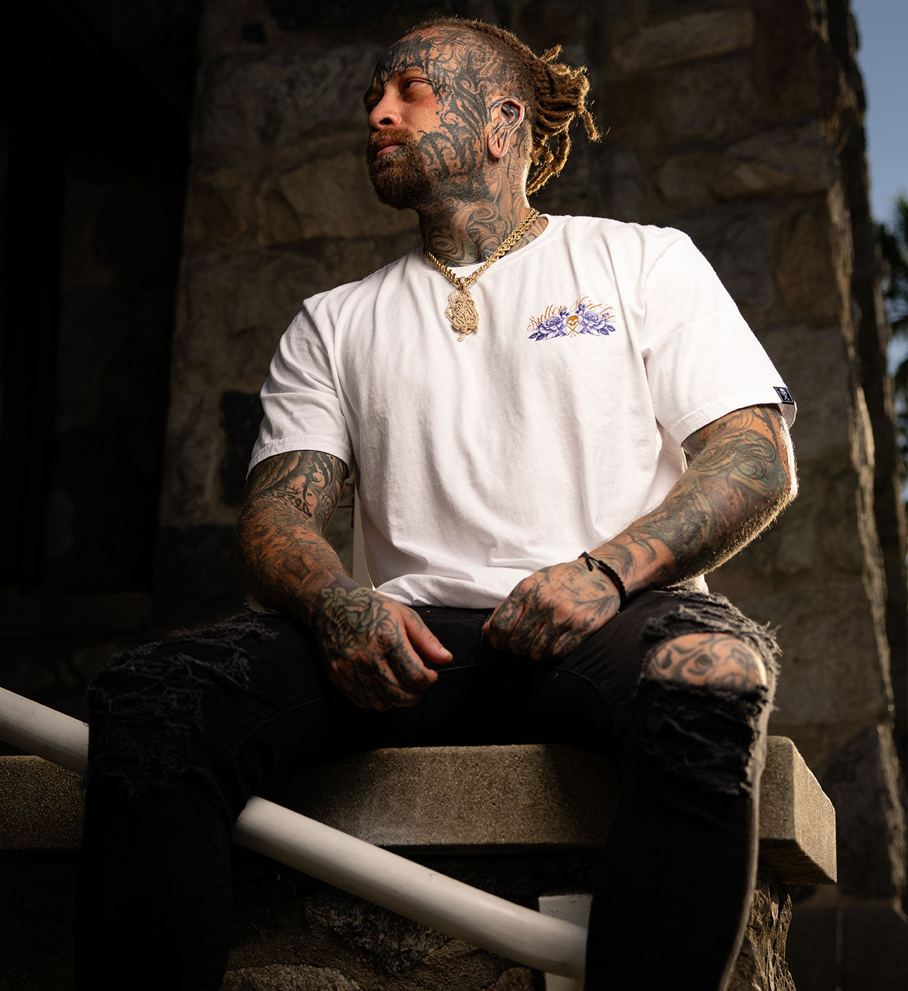 Man with tattoos wearing a white t-shirt with a logo, sitting outdoors.