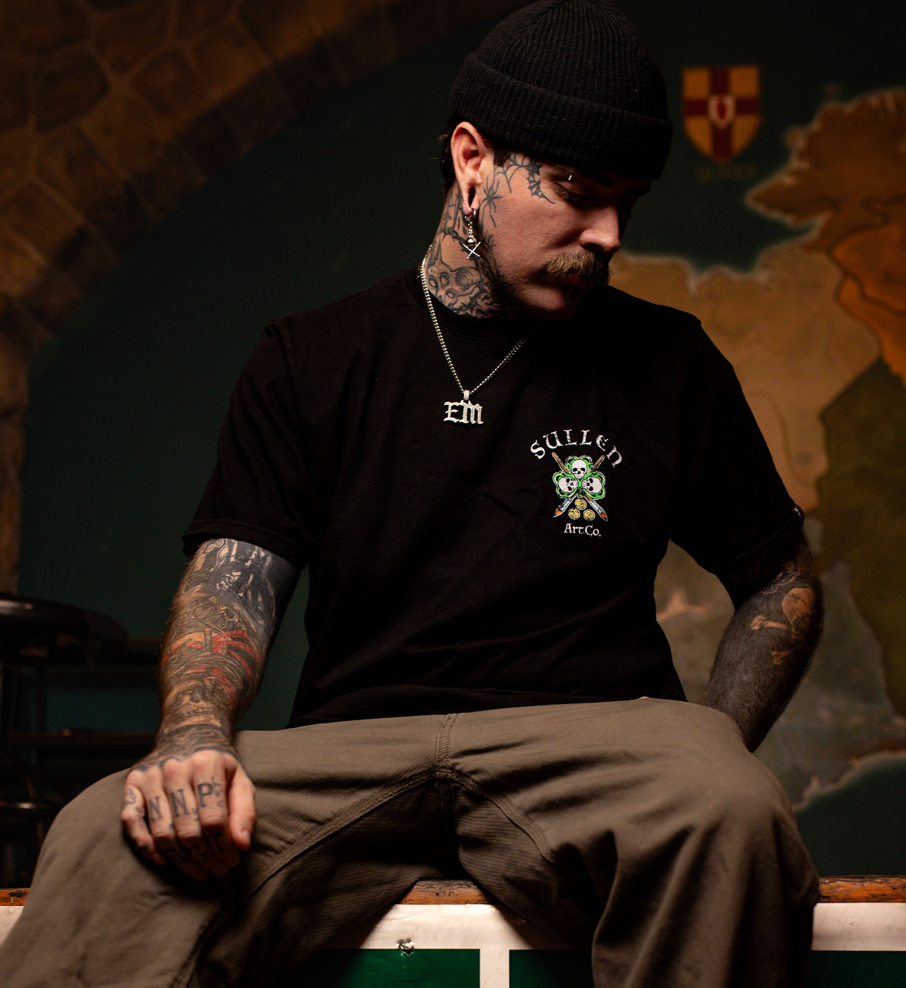 Man wearing a black t-shirt with a logo and text, sitting in a room with a stone wall and coat of arms.