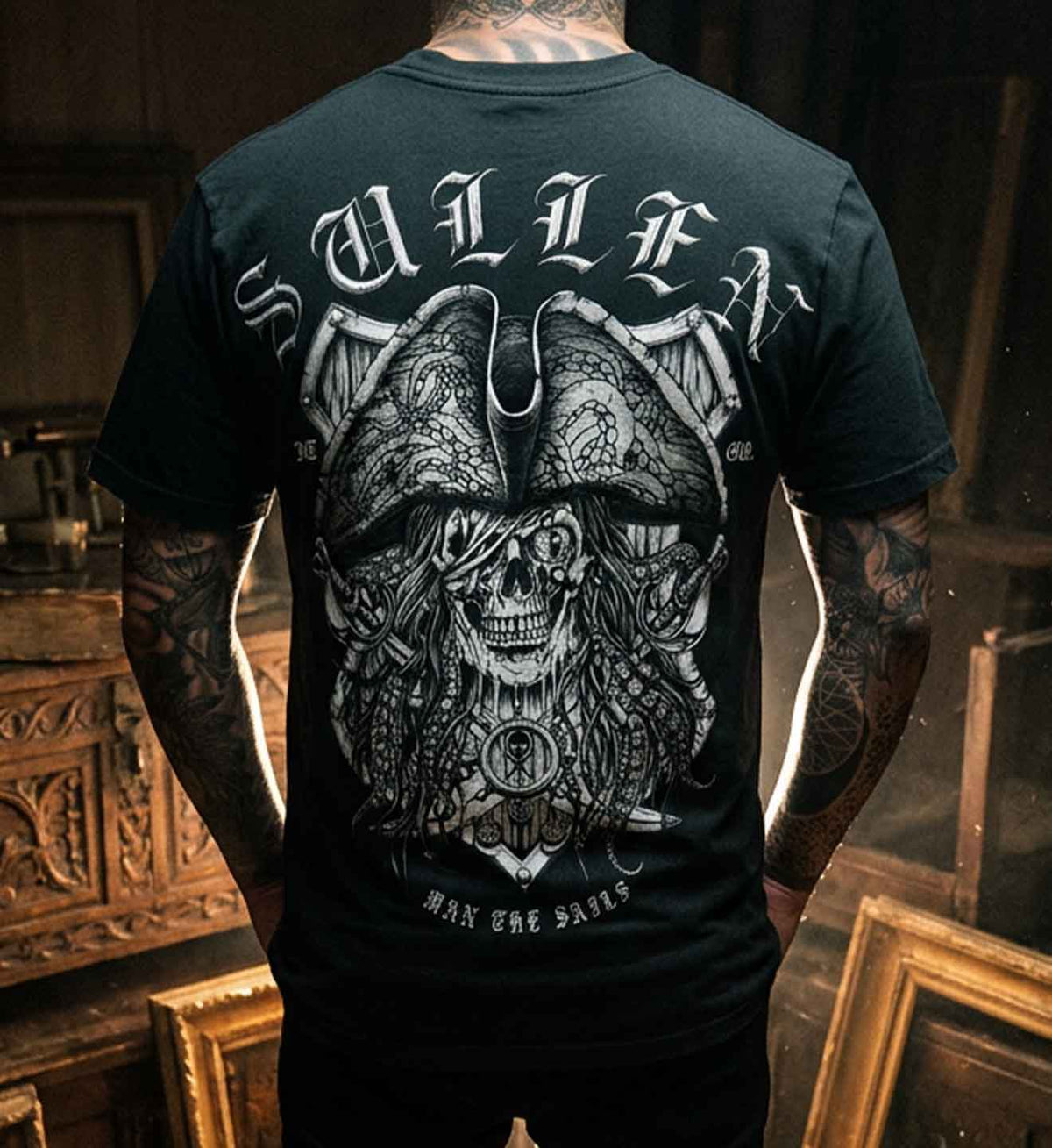 A man viewed from behind wearing a black t-shirt with a large white pirate skull graphic and the word 'SULLEN' across the shoulders, standing in a dark wooden room. Sullen Clothing 2026 Pirate Day capsule tattoo lifestyle dark deep sea treasure hunter artwork.
