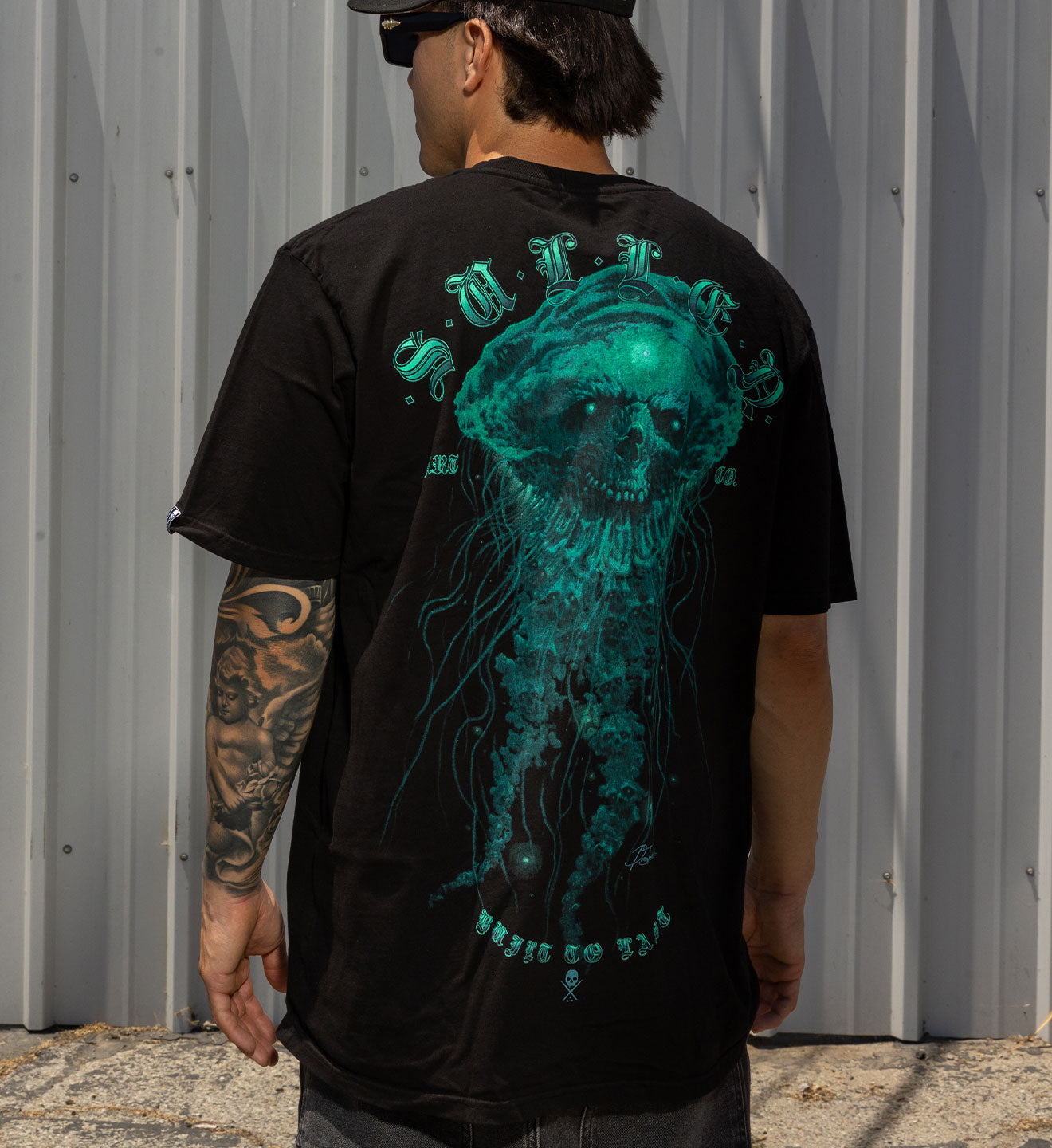 Person wearing a black t-shirt with a teal jellyfish design on the back, standing against a gray wall.