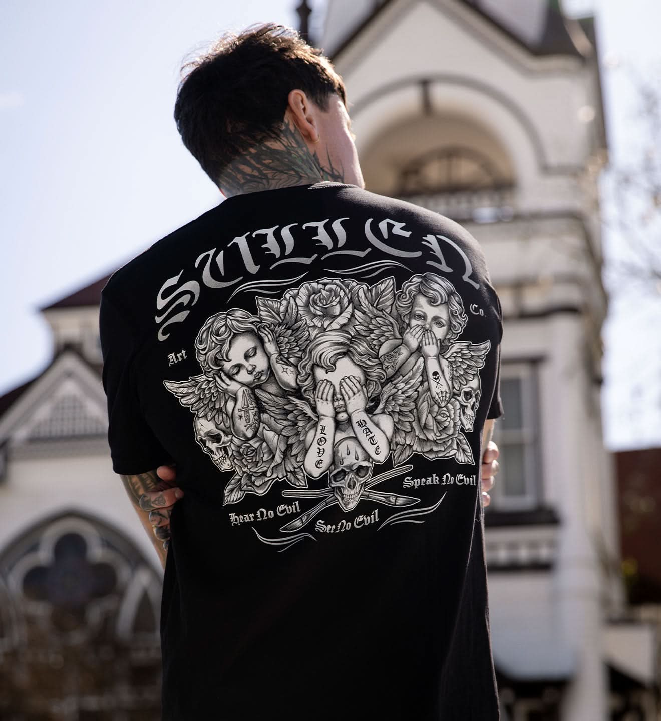 Person wearing a black t-shirt with intricate designs and text, standing in front of an architectural building.