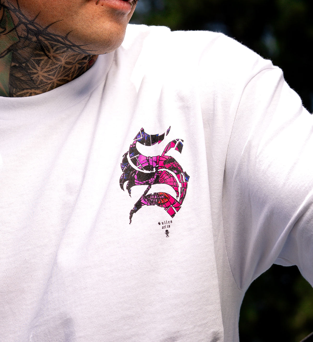 White t-shirt with a colorful graphic design on the chest, worn by a person with tattoos.
