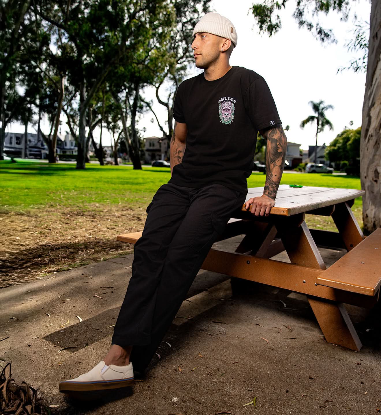 Man sitting on a wooden picnic table in a park wearing a black t-shirt and pants.