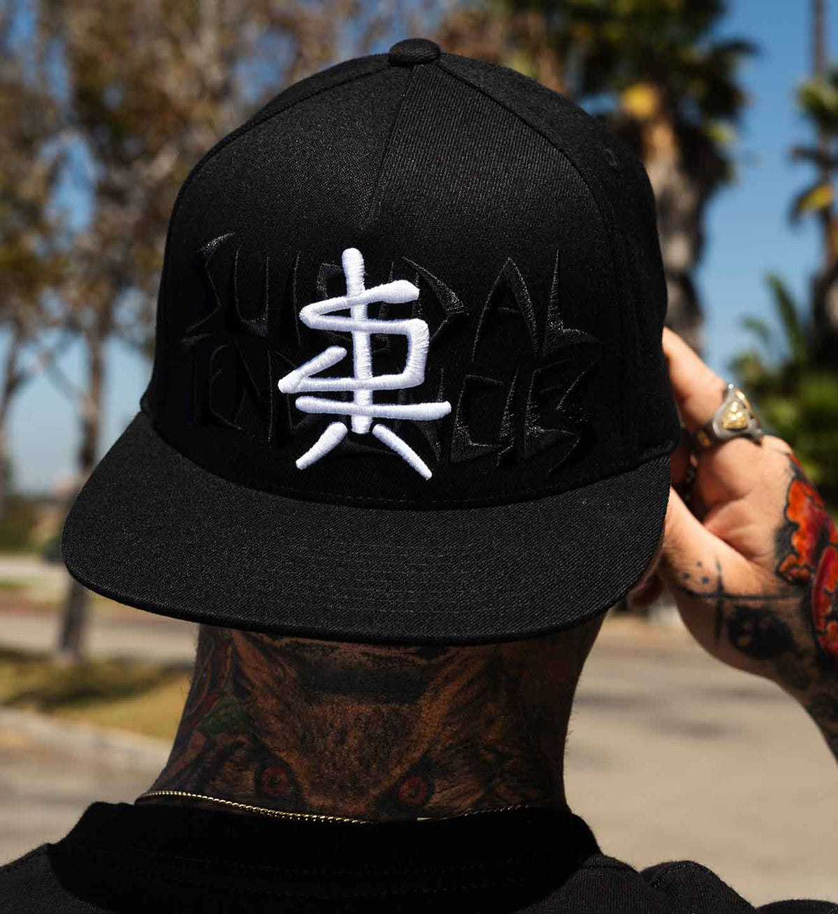 A black flat-brim baseball cap with a large white embroidered Chinese character on the front from Suicidal Tendencies, worn by a tattooed person outdoors with palm trees and a street in the background.