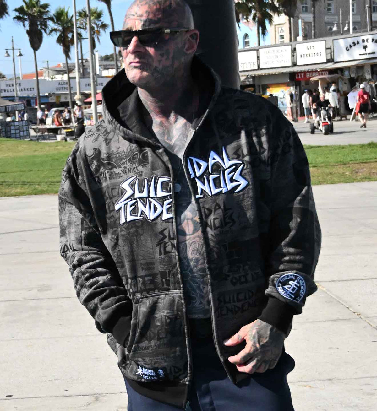 A tattooed man wearing a black graphic zip-up hoodie with 'Suicide Tendencies' text, standing outdoors on a sunny day.