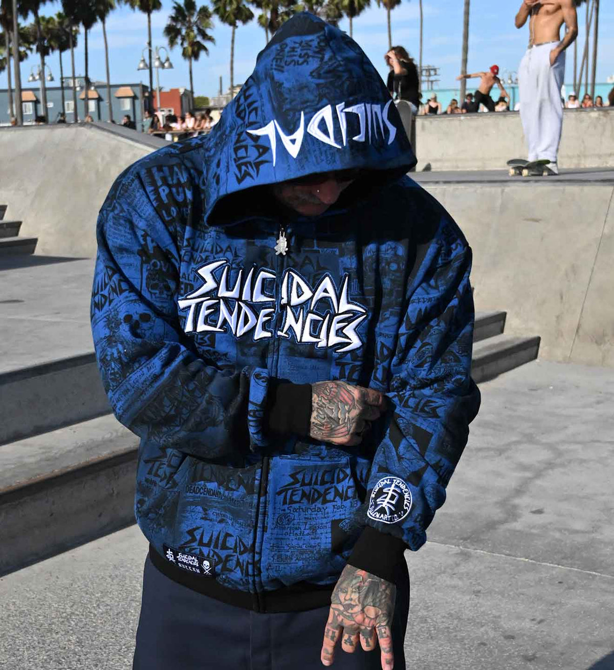 A person wearing a blue zip-up hoodie with 'Suicidal Tendencies' text and graffiti-style graphics, photographed at an outdoor skatepark.