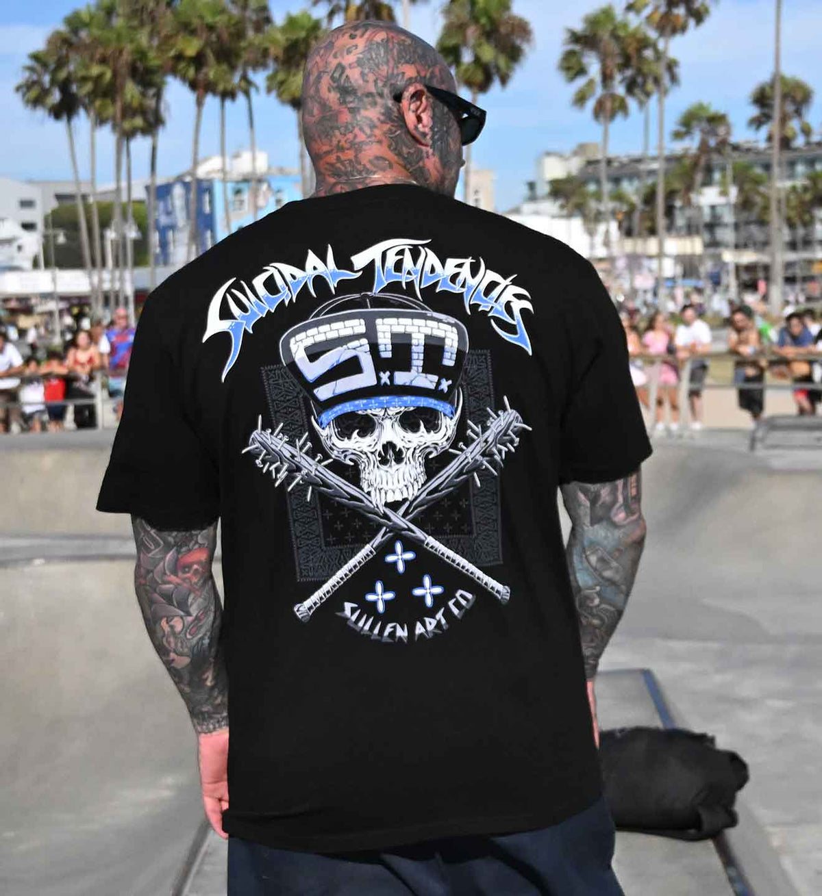 A man viewed from behind wearing a black t-shirt with a large graphic on the back. The graphic features the text 'SUICDIAL TENDENCIES' in a stylized font, a white skull wearing sunglasses, crossed barbed wire, and 'SULLEN' ART CO' at the bottom.