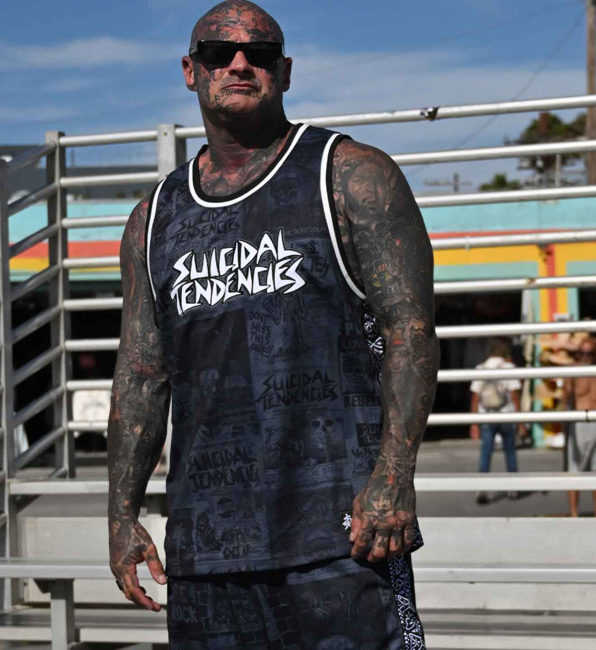 A tattooed man wearing a black graphic tank top with 'SUICIDAL TENDENCIES' printed in white across the chest, standing outdoors in front of metal railings and a colorful building.
