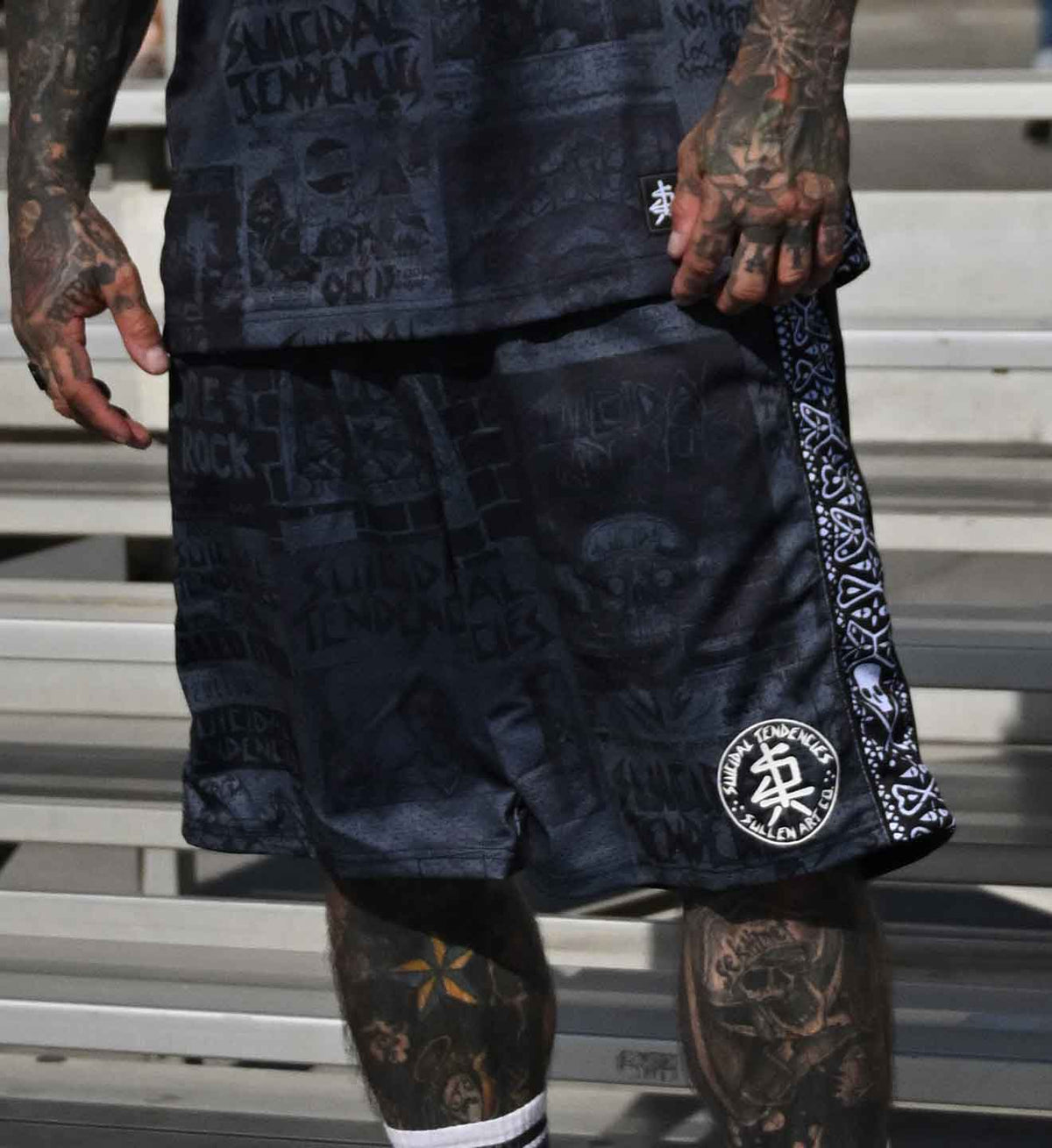 Black graphic athletic shorts with a white bandana print down the side and a circular logo patch near the hem, worn by a tattooed model sitting on metal stadium steps.